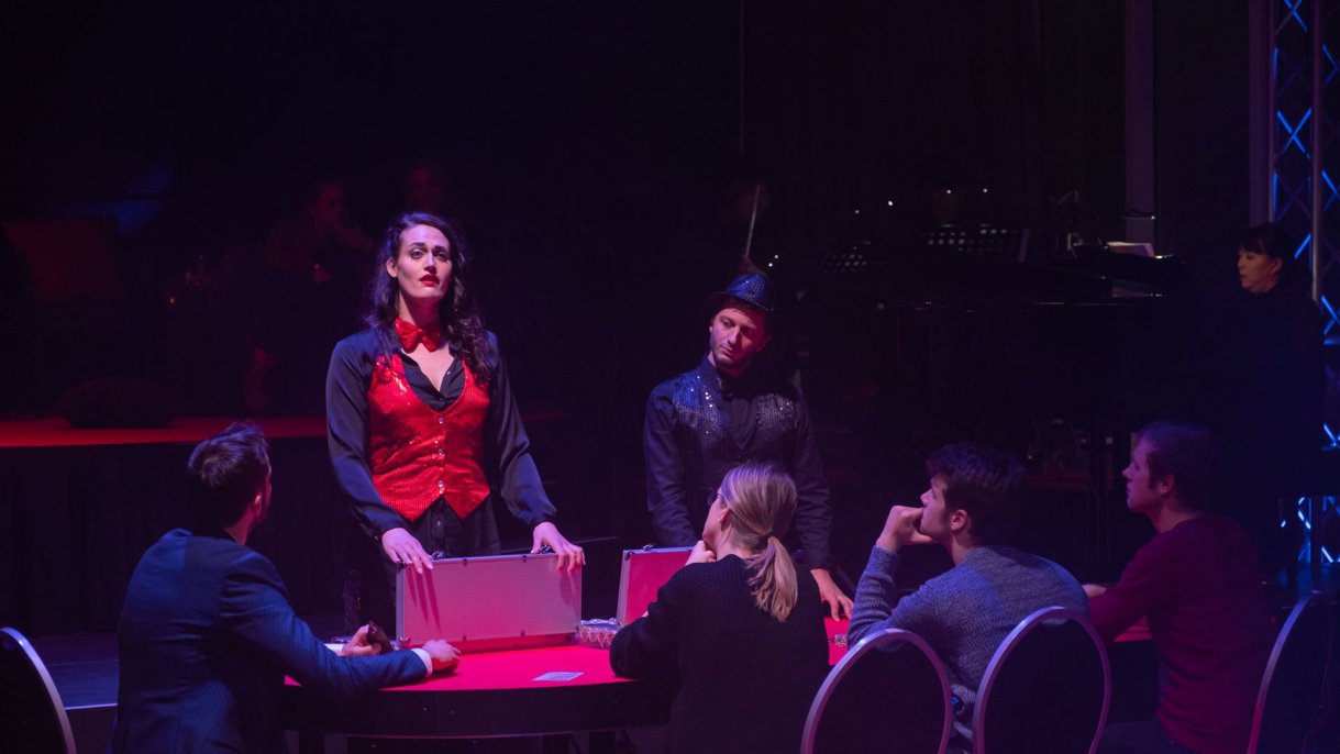 Violetta steht am Pokertisch, an dem Gäste sitzen 