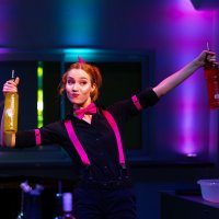 Carmen hält tanzend zwei Flaschen mit bunten Getränken in die Luft, während sie hinter dem Tresen steht.