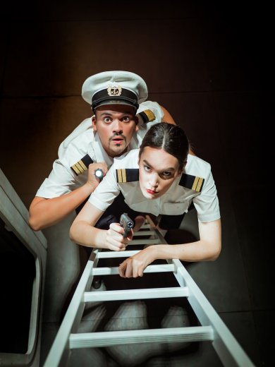 Sängerin und Sänger als Offiziere gekleidet blicken eine Leiter hinauf